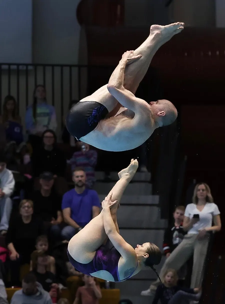 Two divers in a tucked position in mid-air during a synchronized dive, with spectators behind them.